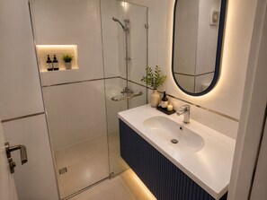 A modern bathroom with a glass shower enclosure, a white sink with a dark vanity, and a round mirror with a blue frame. - MISK Marina Valley Alamein (Alamein)