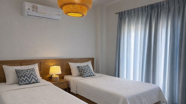 Hotelkamer met twee bedden, een bedlampje, een wandairconditioner en een groot raam met gordijnen.