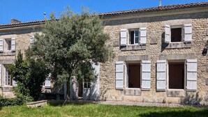 Un bâtiment en pierre doté de nombreuses fenêtres et volets, entouré d’herbe et d’un arbre.