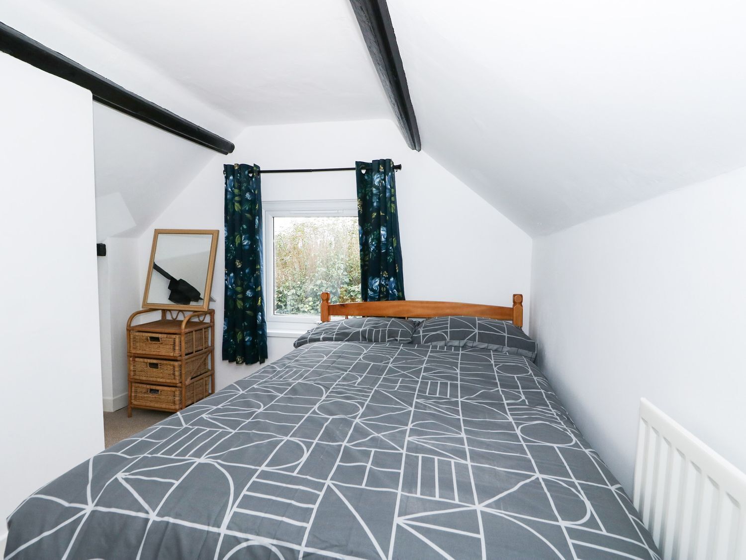 A bedroom with a sloped ceiling, a bed with a geometric-patterned bedspread, a wooden headboard, a wicker nightstand with a mirror, and a window with curtains.