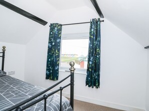 A bedroom with a bed, a window with curtains, a view of buildings outside, and a ceiling with exposed beams. - Oar Cottage (Mablethorpe)