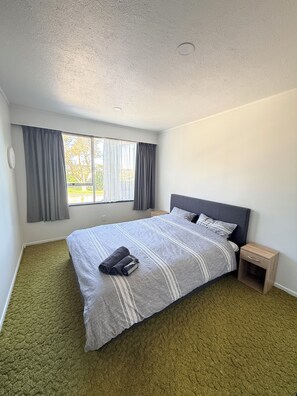A bedroom with a large bed, a nightstand, a window with curtains, and a carpeted floor. - Virginia Lake Family Retreat (Whanganui)