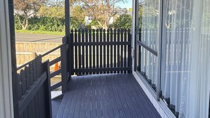 Een overdekt terras met een houten leuning, uitzicht op bomen en een hek, en een heldere lucht.