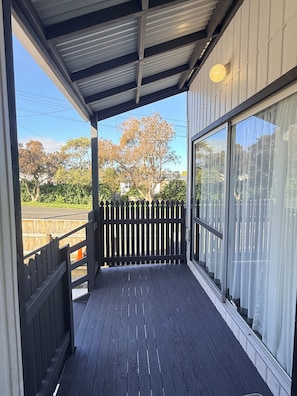 A covered porch with a wooden railing, a view of trees and a fence, and a clear sky.