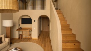 Uma sala de estar ampla com escada de madeira que dá para um andar superior, uma área de jantar com mesa e cadeiras, e um espaço de estar aconchegante com luminária e almofadas.