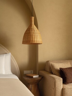 A bedroom with a bed, a bedside table with a book, a brown woven pendant light, and a brown armchair.