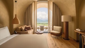 Quarto de hotel com uma cama, um sofá, uma cadeira, uma mesa e um abajur. É possível ver o exterior por uma janela grande.