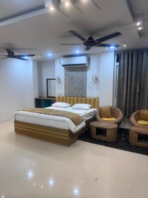 A modern bedroom with a large bed, two wall-mounted fans, an air conditioning unit, and a seating area with wicker chairs. - Hotel Krishna & Restaurant (Barharwa)