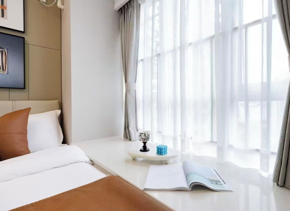 Hotelkamer met een bed, nachtkastje met een boek en decoratie, een raam met dunne gordijnen en uitzicht op de stad.