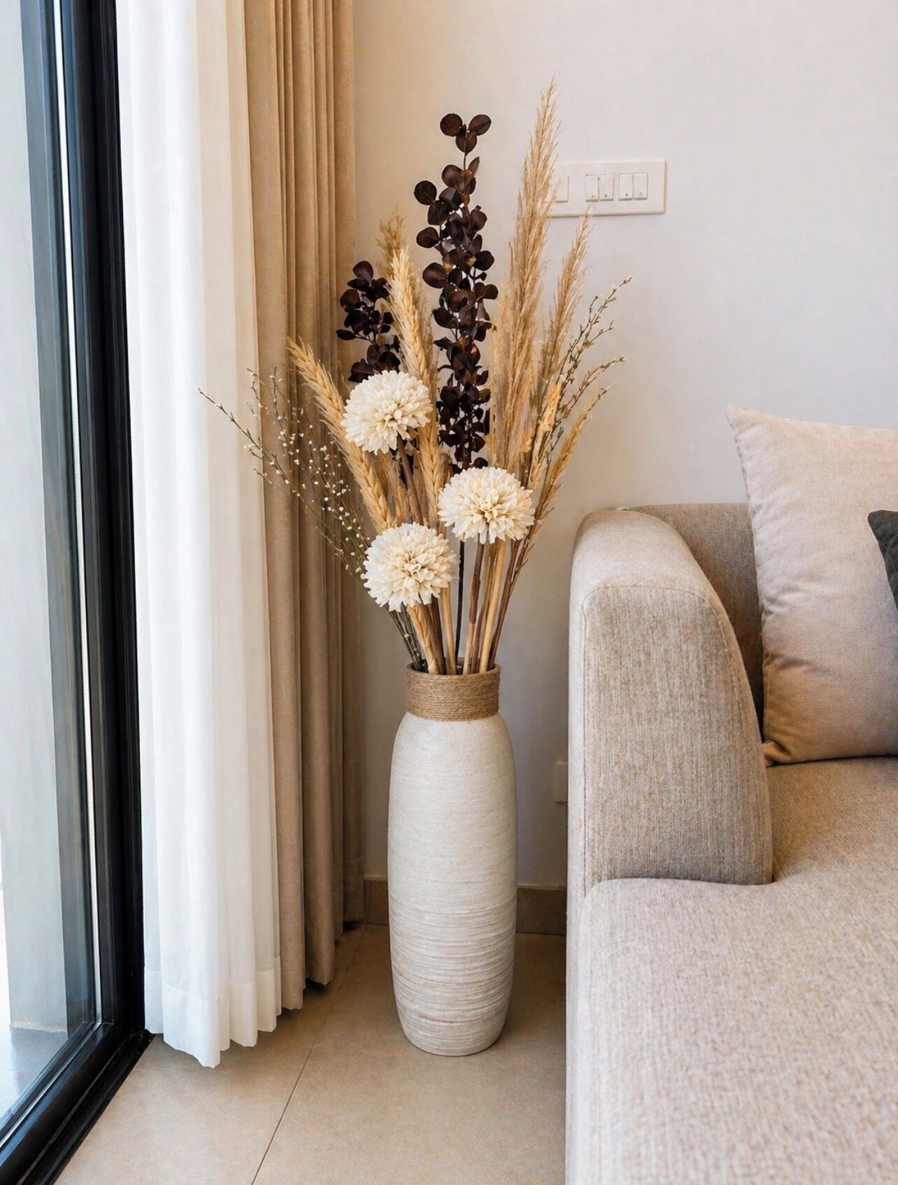 Une pièce avec un canapé beige, un grand vase contenant des plantes séchées et une fenêtre avec des rideaux.