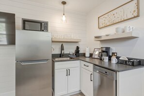 A modern kitchen with a stainless steel refrigerator, microwave, and dishwasher. There are white cabinets, a sink, and a coffee bar with mugs and a coffee maker. - Birch @ Timberline Flats (Traverse City)
