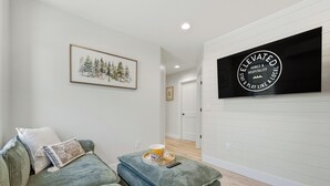 A living room with a sofa, ottoman, framed artwork, and a mounted television. - Birch @ Timberline Flats (Traverse City)