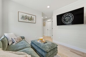 A living room with a sofa, ottoman, framed artwork, and a mounted television. - Birch @ Timberline Flats (Traverse City)