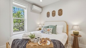 A bedroom with a bed, a window, a wall-mounted air conditioner, two wall art pieces, a bedside lamp, and a tray with pastries and a plant. - Birch @ Timberline Flats (Traverse City)