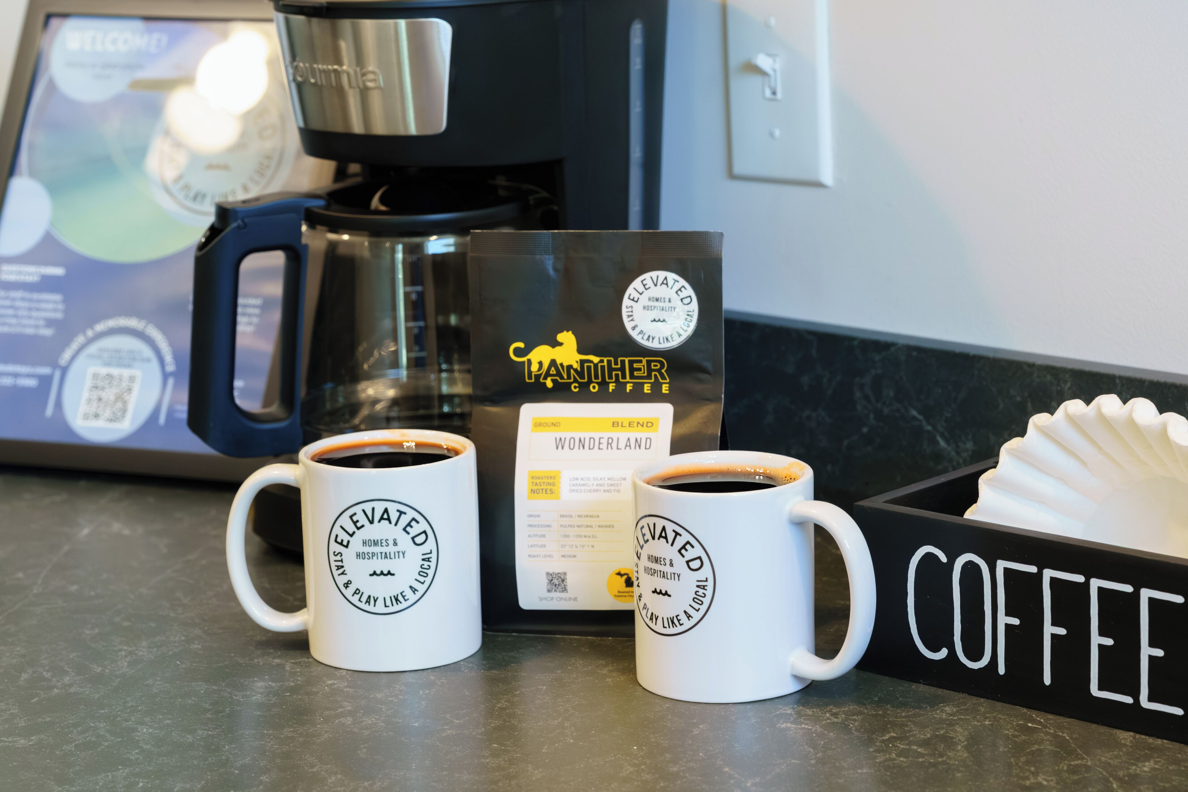 Deux tasses blanches ornées du logo « Elevated », une machine à café et une boîte étiquetée « Panther Coffee Wonderland ».