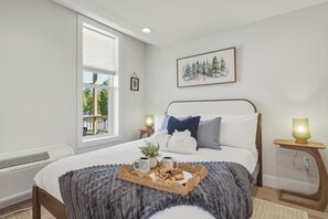 A bedroom with a bed, a window, a framed picture, bedside tables, and a tray with food and drinks. - Timberline 4 upper Units (Traverse City)