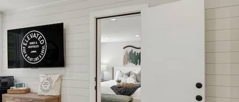 A modern hotel room with a flat-screen TV, a wooden side table, a bed with a green upholstered bench, and a white door.