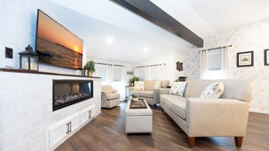 A living room with a fireplace, a sofa, a coffee table, and a TV mounted on the wall. - Sirens Keep | Boutique Style Outdoor Living • Hot Tub • Sleeps 8 (Delhaven)