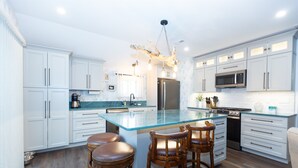 A modern kitchen with white cabinetry, a central island with stools, stainless steel appliances, and a chandelier. - Sirens Keep | Boutique Style Outdoor Living • Hot Tub • Sleeps 8 (Delhaven)