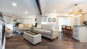 A modern living room with a sofa, ottoman, and a dining area with chairs and a table. - Sirens Keep | Boutique Style Outdoor Living • Hot Tub • Sleeps 8 (Delhaven)
