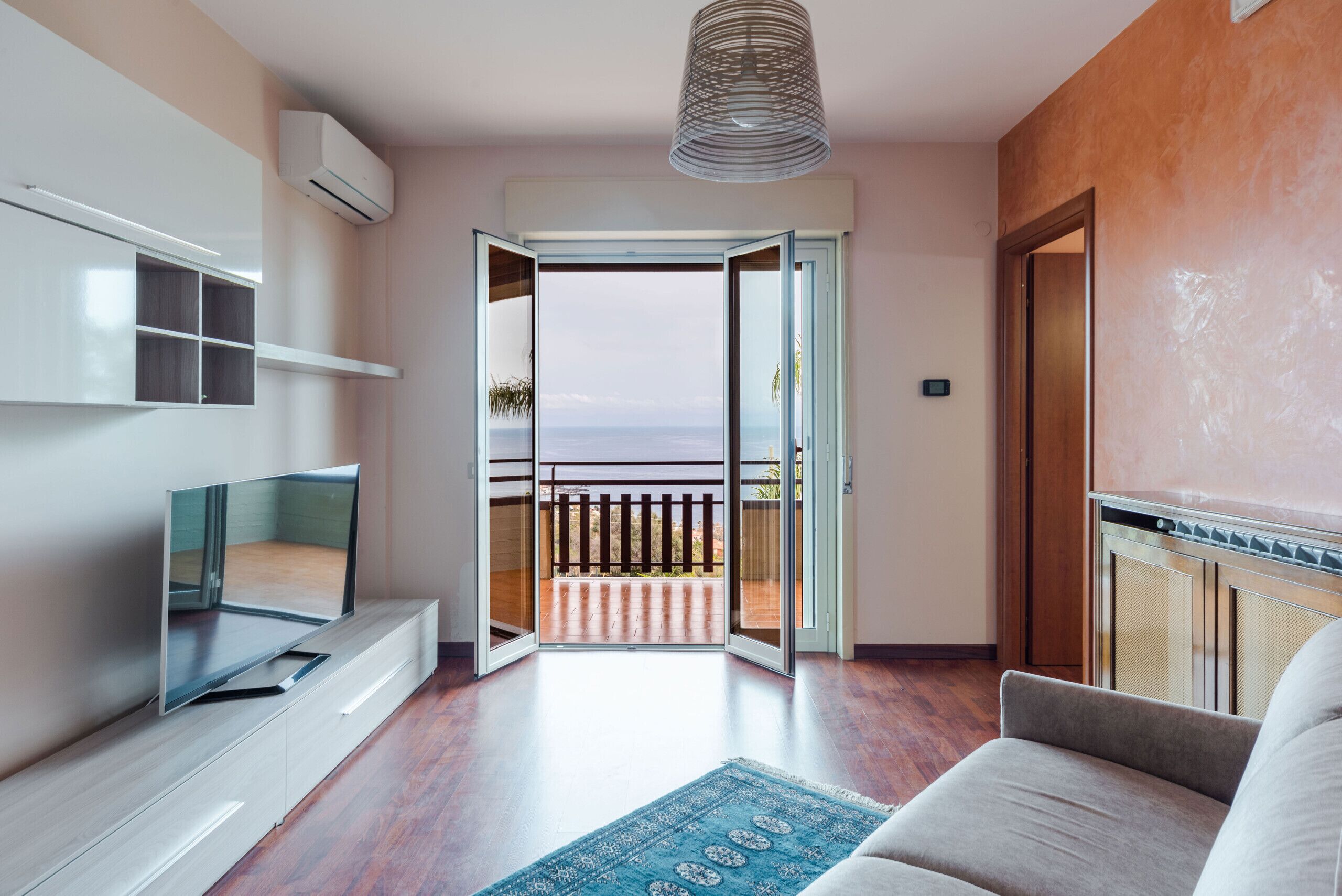 Ein modernes Wohnzimmer mit Fernseher, einer Couch, einem gemusterten Teppich und Blick aufs Meer durch eine offene Tür.