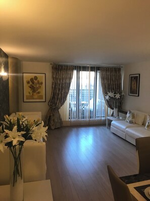 Een woonkamer met een bank, een salontafel, een stoel, een vaas met bloemen en een groot raam met gordijnen.