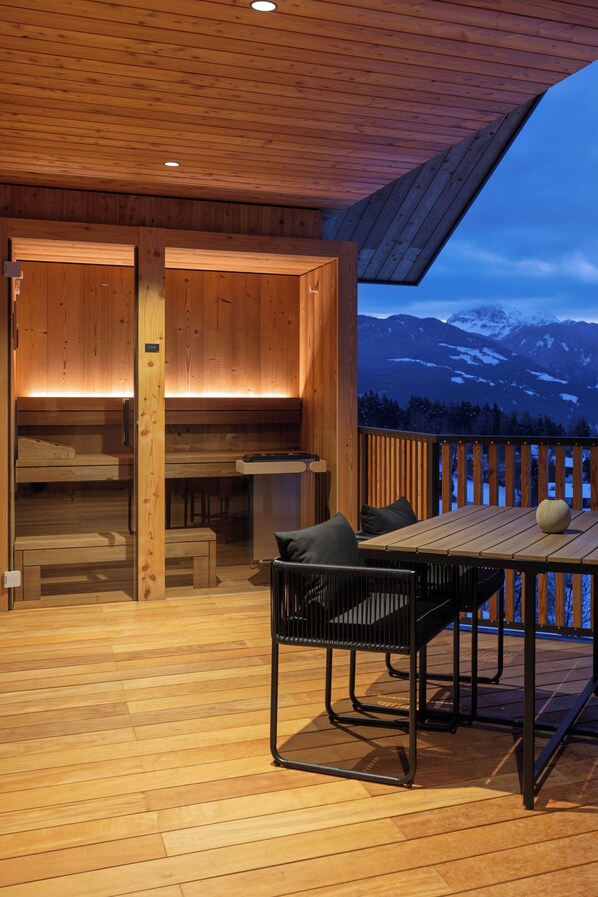A wooden deck with a table and chairs, a view of mountains, and a wooden interior with benches. - Oachner Living | Design vacation apartments with sauna in South Tyrol (Trentino-Alto Adige)