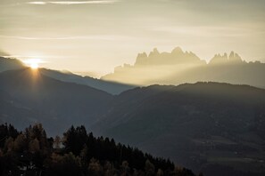 A mountainous landscape with a house in the foreground and a sunlit mountain range in the background. - Oachner Living | Design vacation apartments with sauna in South Tyrol (Trentino-Alto Adige)