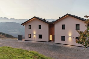 A modern two-story building with a symmetrical facade, large windows, and a gravel driveway. - Oachner Living | Design vacation apartments with sauna in South Tyrol (Trentino-Alto Adige)