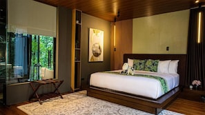 A modern bedroom with a large bed, a wooden headboard, a bedside table, a nightstand, a window with a view of greenery, and a wall-mounted artwork. - House of Gaia (Lipa City)