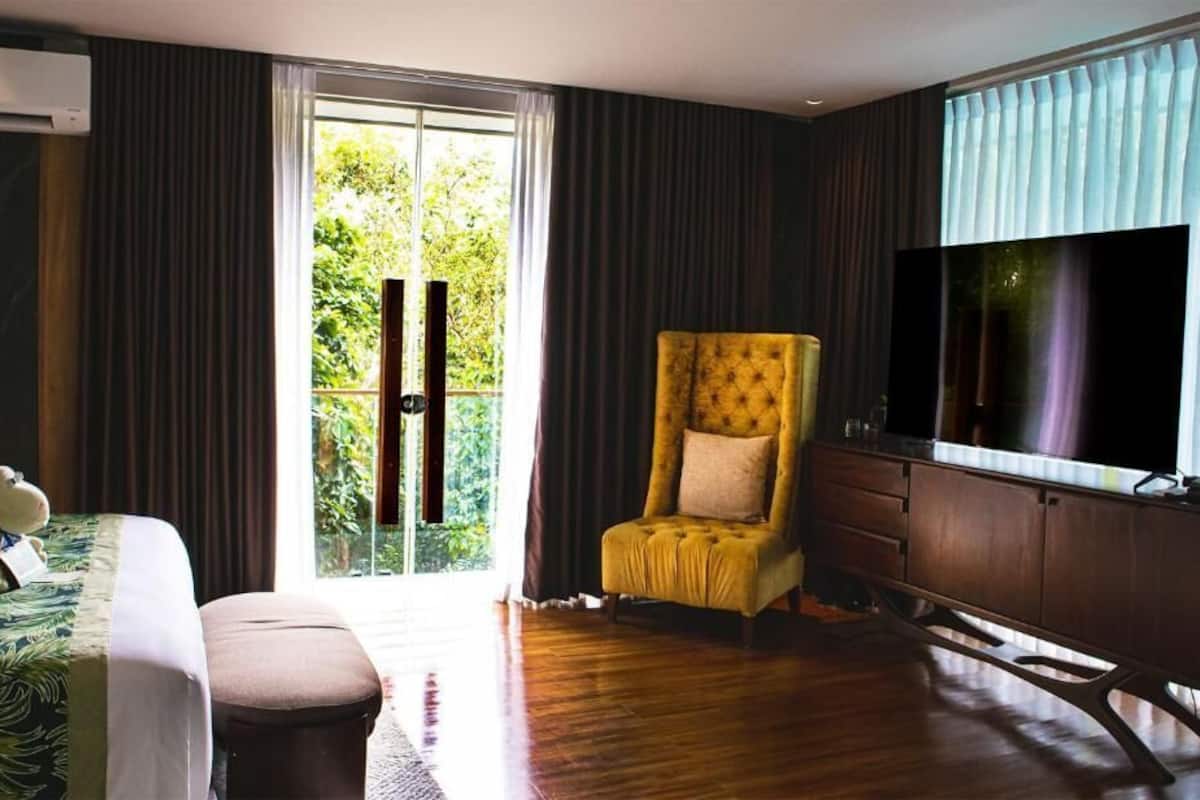 Habitación de hotel con cama, una silla, escritorio, televisor y puerta corrediza de vidrio a un balcón.