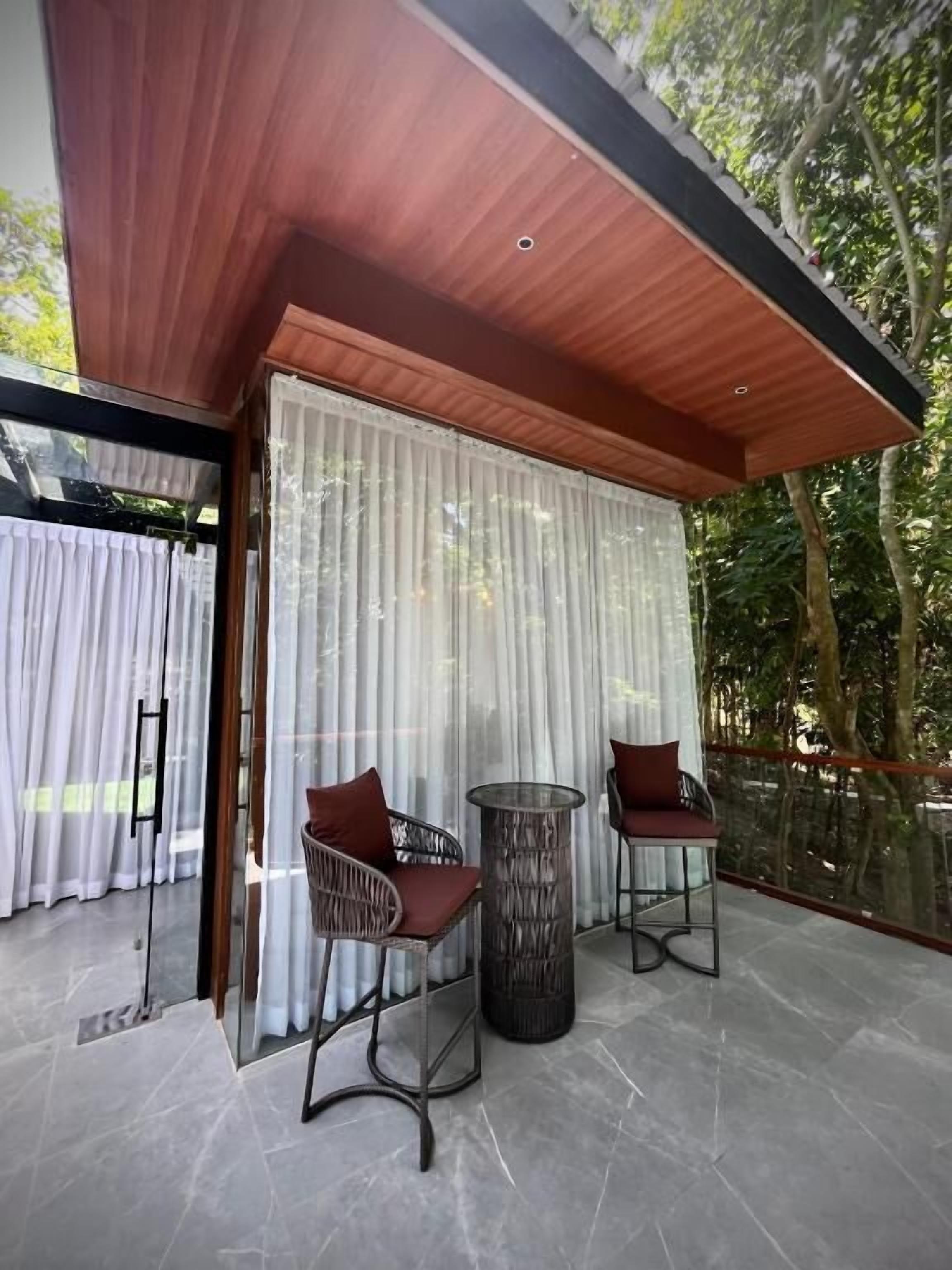 A modern outdoor seating area with two wicker chairs and a round table, surrounded by a glass railing and a wooden ceiling.