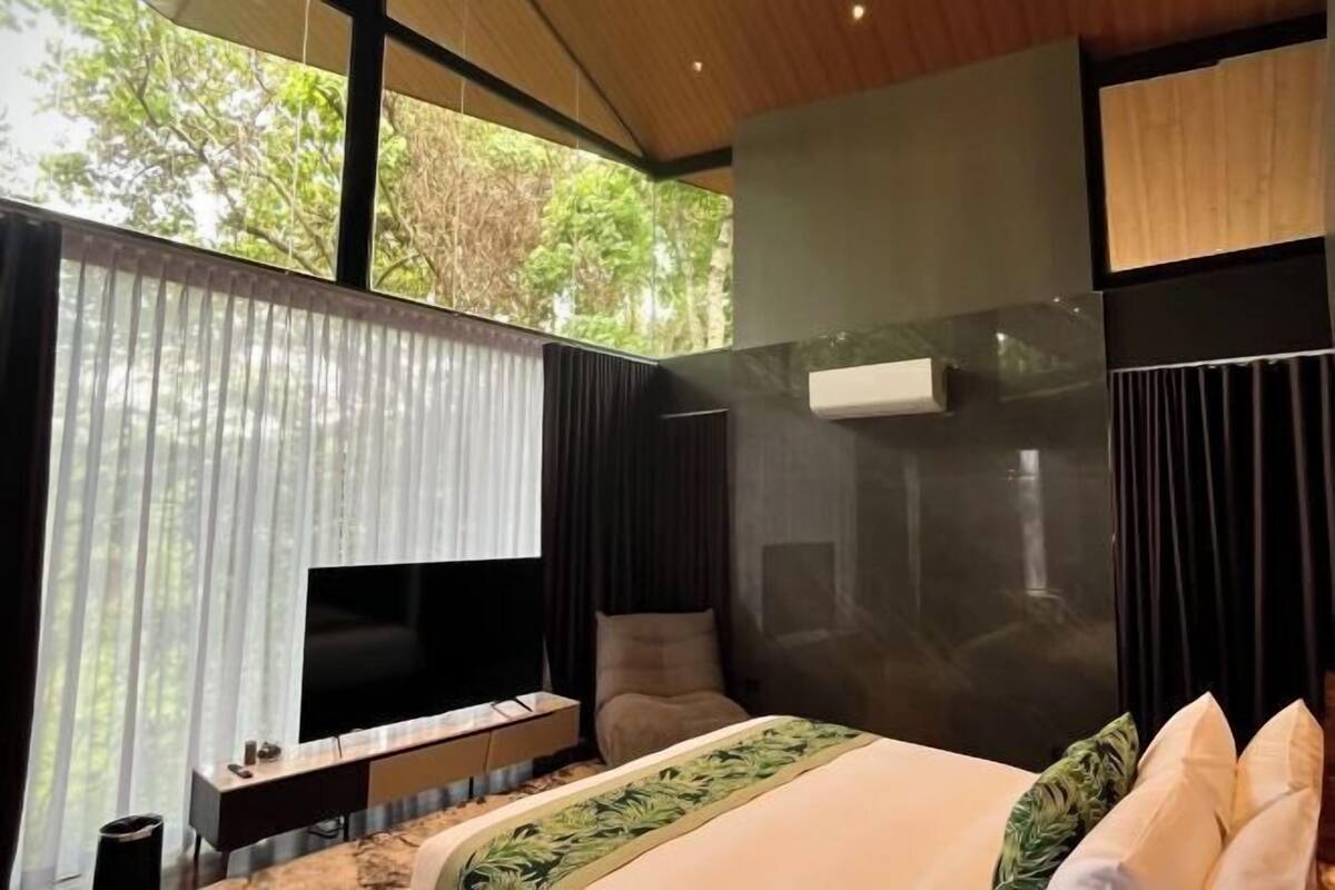 Un dormitorio moderno con una cama grande, un televisor montado en la pared y vistas a árboles por la ventana.