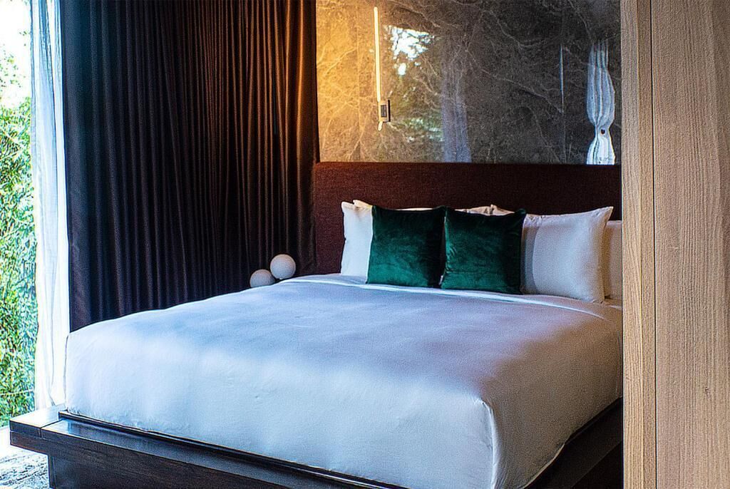 A modern bedroom with a large bed, dark curtains, and a view of trees through the window.