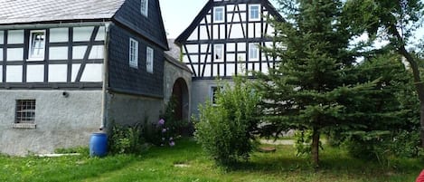A traditional half-timbered house with a sloped roof, a small garden with a tree, and a blue barrel.