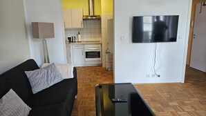 Sala de estar con un sofá negro, una mesa de centro de vidrio, un televisor de pantalla plana montado en la pared y una cocina visible a través de un vano abierto.