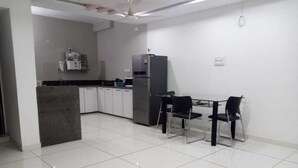 A modern kitchen with white cabinets, a refrigerator, and a dining area with two chairs and a glass table. - Hotel nova galaxy  (Rajkot)