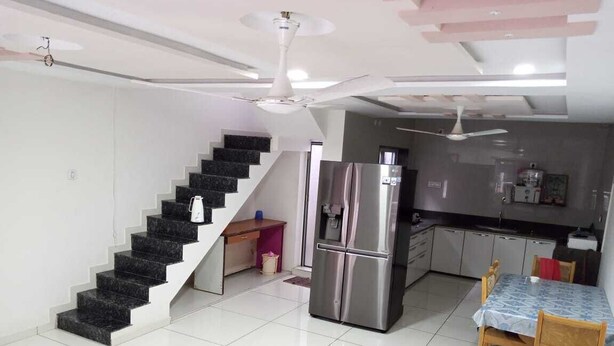 A modern kitchen with a stainless steel refrigerator, a dining table with chairs, and a staircase with a black railing. - Hotel nova galaxy  (Rajkot)