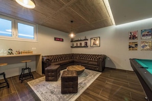 A room with a curved leather sofa, a pool table, a bar area with stools, and a ceiling with exposed beams.