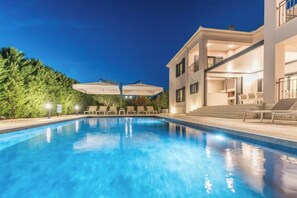 Una piscina con lettini e ombrelloni, un edificio a più piani con balconi e un siepe sullo sfondo.