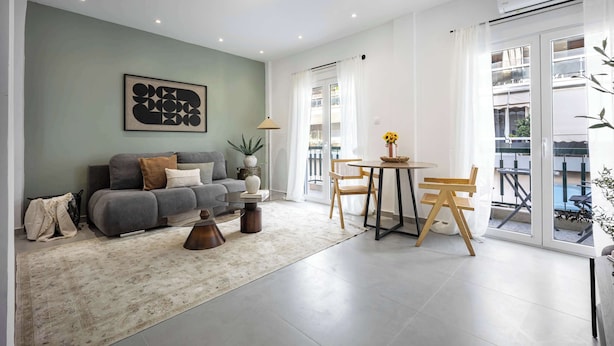 En moderne stue med sofa, sofabord og stole, et spiseområde med bord og stole, samt et stort vindue med udsigt til en balkon.