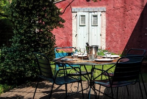Un espace de restauration en plein air avec une table dressĂ©e pour quatre personnes, entourĂ©e de chaises en mĂ©tal, le tout adossĂ© Ă un mur rouge percĂ© dâune fenĂȘtre.