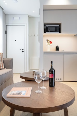 A modern living room with a round wooden table, two wine glasses, a bottle of wine, a magazine, a sofa, and a white door. - Trilogia Suites (Athina)