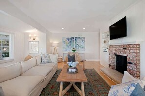 A living room with a sofa, a coffee table, a fireplace, and a television mounted on the wall. - Oak & Ivy Cottage - Heart of Montecito (Santa Barbara)