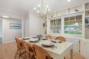 A dining area with a white table set for four, wicker chairs, a chandelier, and a view through a window with a white fence. - Oak & Ivy Cottage - Heart of Montecito (Santa Barbara)