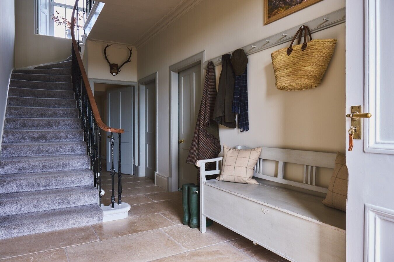 A staircase with a wooden handrail, a wall-mounted deer head, a bench with a woven bag, and a door with a brass handle.