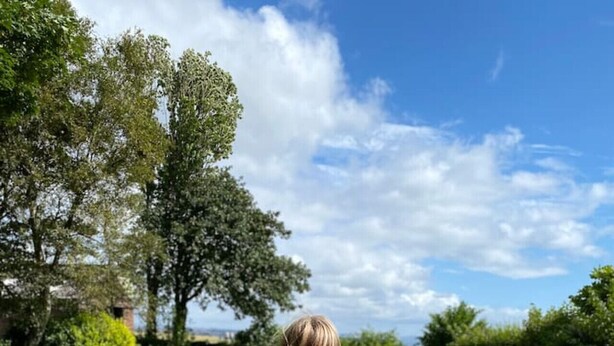 Een persoon zit op een deken in een grasveld, met een hond en een picknickopstelling, inclusief een mand, flessen en eten.