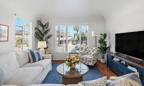 A living room with a sofa, armchair, coffee table, TV, and large windows. - The Palm Terrace - West Beach Retreat (Santa Barbara)
