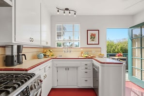 A kitchen with white cabinets, a gas stove, a coffee maker, and a window with blinds. - The Palm Terrace - West Beach Retreat (Santa Barbara)