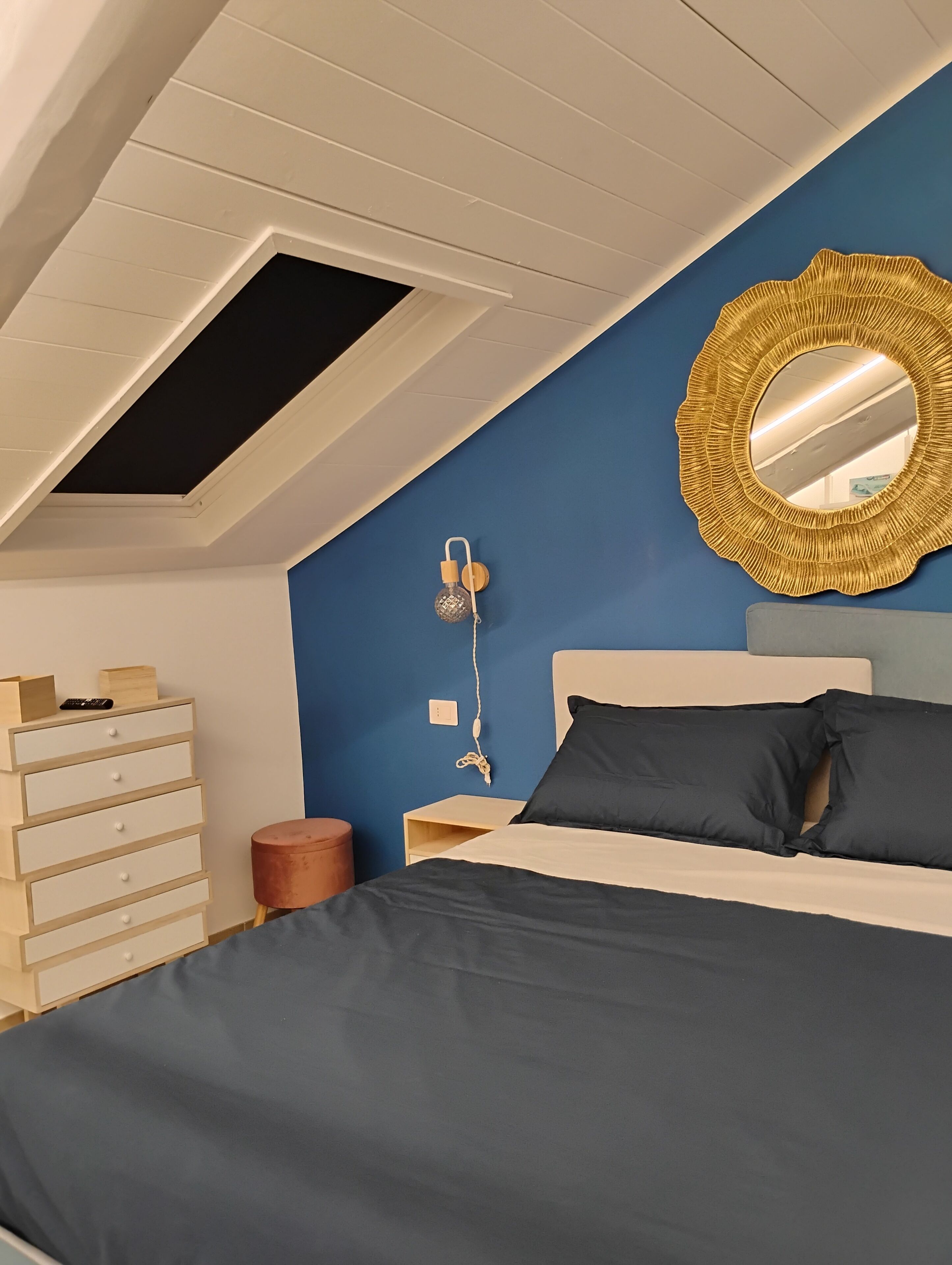 Une chambre avec un plafond en pente, un lit avec une literie aux couleurs sombres, une commode en bois, un miroir rond et une lampe fixée au mur.